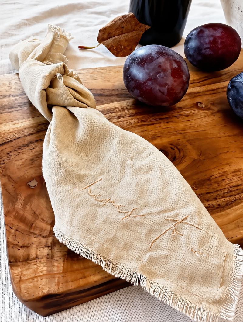 Buttercream linen napkin with fringe edge on a wooden board beside plums